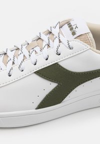 Sneaker bianco con un accento di camoscio verde oliva, linguetta beige texturizzata e lacci bianchi. Materiale liscio con dettagli perforati sul lato.