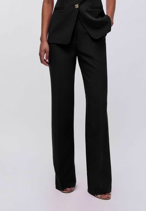 Pantalones negros de talle alto y pierna ancha, combinados con un blazer negro y tacones abiertos, modelo de pie con una mano en el bolsillo.