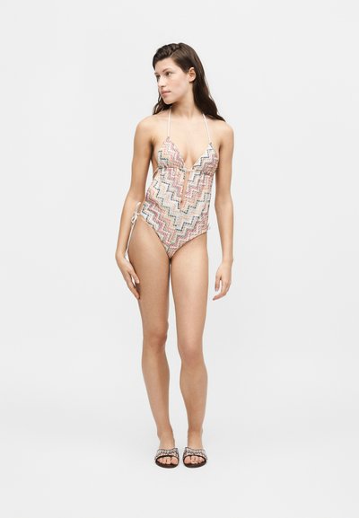 Νεαρή γυναίκα που φοράει πολύχρωμο ολόσωμο μαγιό με ζιγκ-ζαγκ μοτίβο και κορδόνια που δένουν στον λαιμό, καθώς και αντιστοιχισμένα σανδάλια σε απλό φόντο.