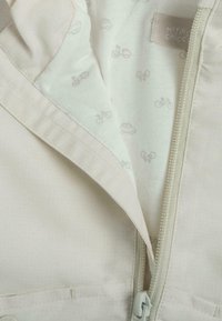 Veste légère beige avec une fermeture à glissière, doublée d'un tissu vert clair doux, ornée de motifs de véhicules gris clair. L'étiquette indique "MON PREMIER CHICCO."