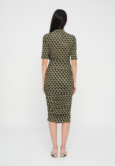 Kobieta z długimi ciemnymi włosami w dopasowanej sukience do kolan w zielono-beżowy wzór w romby, stojąca tyłem na białym tle.