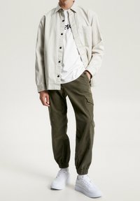 Vaaleanbeige nappipaita valkoisen graafisen t-paidan päällä, yhdistettynä oliivinvihreisiin kapeneviin cargo-housuihin ja valkoisiin lenkkareihin.