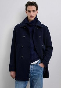 Marineblå uldfrakke med en struktureret, kunstpelskrave, dobbeltbrystet design, sidelommer og en afslappet pasform, båret over en rillet turtle neck.