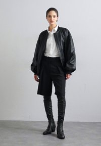 Black leather bomber jacket with a relaxed fit, over a white button-up shirt, paired with tailored black shorts and knee-high black boots.