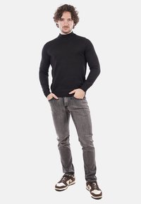 Maglione nero a collo alto, design aderente, abbinato a jeans grigi. Indossato con sneakers marroni e bianche, in piedi su uno sfondo bianco.