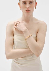 Pendientes de aro en tono dorado, un anillo texturizado y una delicada pulsera de cadena complementan una prenda sin mangas de color crema con una textura suave.