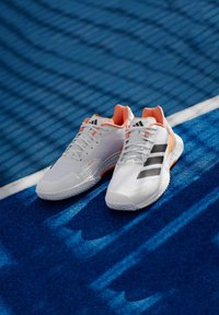 Λευκά αθλητικά παπούτσια με υφή, μαύρες ρίγες και πορτοκαλί λεπτομέρειες σε μπλε γήπεδο. Ελαφρύς σχεδιασμός με καουτσούκ σόλα.