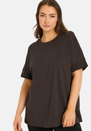 Ung kvinde med langt brunt hår iført en løs sort kortærmet skjorte, stående mod en ensfarvet lys baggrund.