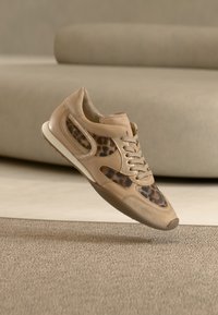 Μπεζ αθλητικό παπούτσι με λεπτομέρειες λεοπάρ επιπλέει πάνω από χαλί με υφή σε μπεζ χρώμα μέσα σε δωμάτιο με ουδέτερο τόνο και με καμπυλωτό καναπέ στο βάθος.