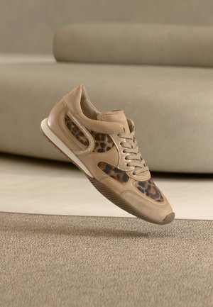 Beige värinen tennari, jossa leopardikuvioiset yksityiskohdat, leijuu teksturoidun beigen sävyisen maton yllä neutraaleissa sävyissä olevassa huoneessa, taustalla kaareva sohva.