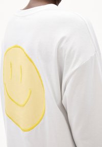 Camicia bianca con un grande patch ricamato con una faccina sorridente gialla sulla parte superiore della schiena, indossata da una persona vista da dietro.