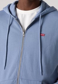Felpa con zip di colore azzurro chiaro, dotata di cordini, polsini a coste e un piccolo logo rosso Levi's sul lato sinistro del petto. Il tessuto è morbido e a trama.