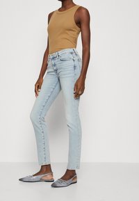 Jeans de tiro alto en azul claro combinados con una blusa sin mangas color beige y zapatos de tacón bajo en gris, con punta afilada y detalle de hebilla.