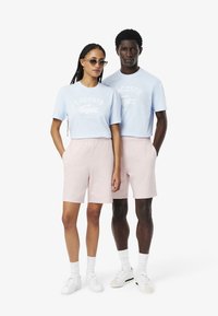 Due modelli indossano outfit coordinati: t-shirt di cotone azzurro chiaro con logo Lacoste e pantaloni corti rosa. Sono abbinati a sneaker bianche.