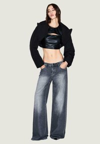Chaqueta negra corta con hombreras acolchadas sobre un top corto de cuero negro ajustado, combinado con jeans anchos de pierna amplia en denim gris desgastado.