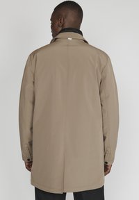 Abrigo impermeable beige con corte recto, diseño minimalista y puños abotonados. Textura suave y sutiles detalles de costura.
