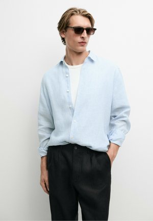 Junger Mann mit dunkler Sonnenbrille, einem hellblauen Hemd über einem weißen T-Shirt und schwarzen Hosen, eine Hand in der Tasche.