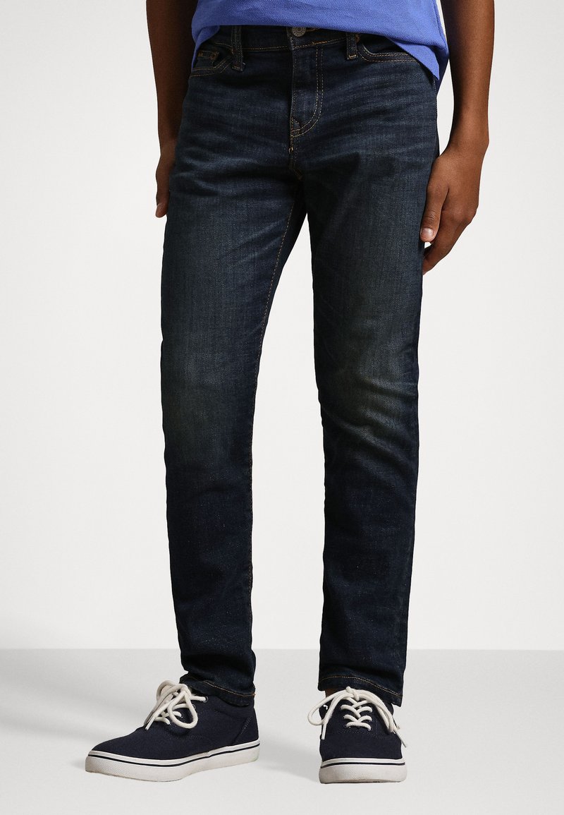 Jeans de ganga azul-escuro com corte slim, apresentando desbotamento subtil, costura laranja e design clássico de cinco bolsos. Acompanhados por sapatilhas azul-marinho.