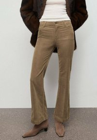 Pantalones de pana color mostaza con corte acampanado, bolsillos delanteros, cremallera, y una sutil textura, combinados con botas de tacón alto color marrón claro.
