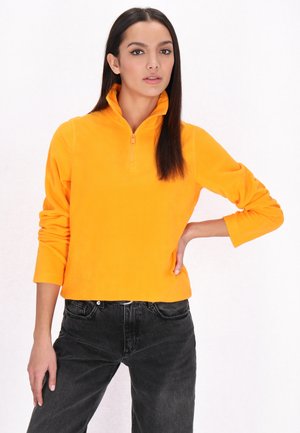 Lys orange fleece pullover med halv-zip krave, lange ærmer og en afslappet pasform, kombineret med sorte jeans på en neutral baggrund.