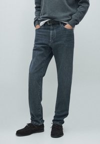 Ciemnoniebieskie dżinsy o prostych nogawkach, z widocznym przeszyciem, pięcioma kieszeniami, noszone z szarym swetrem i czarnymi butami.