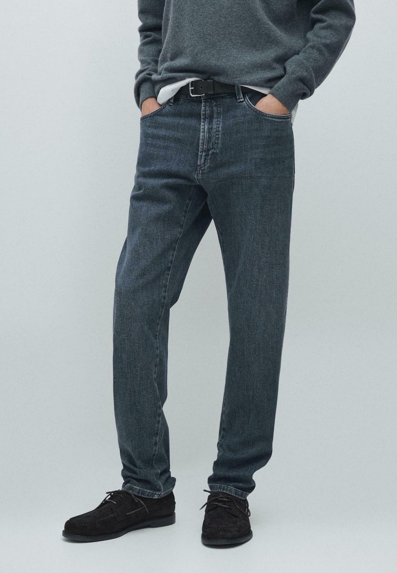 Mørkeblå denimjeans med lige ben, der har synlige syninger, fem lommer og bæres med en grå sweater og sorte sko.