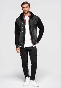 Giubbotto di denim nero con cappuccio nero, indossato sopra una camicia a quadri. Abbinato a pantaloni cargo neri e sneakers bianche. Design semplice e funzionale.