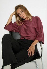 Burgundská oversized halenka s širokými rukávy, strukturovaná tkanina. V kombinaci s černými kalhotami a stříbrným náramkem, sedící na černé židli.