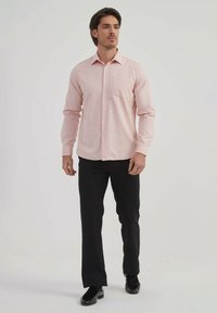 Chemise boutonnée à manches longues rose clair avec une poche unique, associée à un pantalon noir et des chaussures sombres. Tissu lisse et design ajusté.