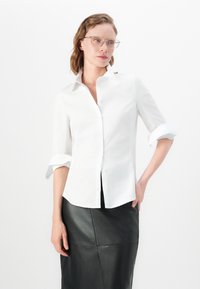 Camicia bianca con maniche arrotolate, realizzata in morbido tessuto di cotone. Indossata con una gonna nera in pelle, caratterizzata da dettagli delle cuciture.