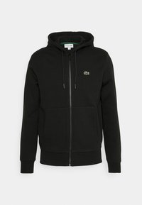 Svart zip-up hoodie i mjuk bomull, med en framficka och broderad grön Lacoste-logotyp på vänster bröst.