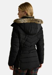 Melnā puffera jaka ar mākslīgā kažokāda apmali kapucē, quiltēta dizaina, ar jostu viduklī un uzsvērta ar zeltītu furnitūru. Redzamas plaid bikses zemāk.