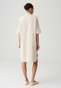 Beiges knielanges Kleid mit Kragen, kurzen Ärmeln, seitlichen Schlitzen und glatter Stoffoberfläche. Kombiniert mit hellen Slippern.