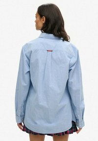 Jasnoniebieska koszula na guziki, wykonana z bawełny, z długimi rękawami, plisą z tyłu i małą czerwoną metką przy kołnierzu.