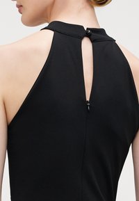 Black sleeveless dress with a high neck, featuring a keyhole back and a black button closure. Smooth fabric with a fitted silhouette.