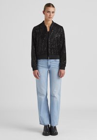 Schwarze Bomberjacke mit Paillettenstruktur, Reißverschluss und gerippten Bündchen und Saum; kombiniert mit hellblauen Jeans im geraden Schnitt und schwarzen Stiefeln.