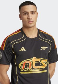 Black sports jersey with red and gold accents, featuring a large logo in orange and a club emblem on the chest. Comfortable fit and fabric.