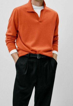 Man wearing a bright orange long-sleeve sweater over a white shirt, tucked into black pleated trousers with hands in pockets.