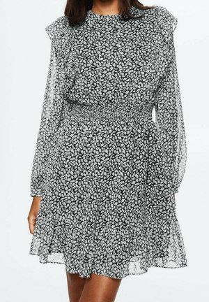 Femme portant une robe fleurie noire et blanche avec des manches longues transparentes, des épaules à volants, une taille cintrée et un ourlet à volants.