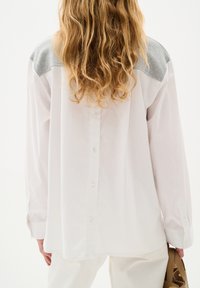 Camicia bianca con inserti in tessuto grigio sulle spalle, maniche lunghe e vestibilità ampia. Chiusura con bottoni e orlo arrotondato dettagliato ai polsini.