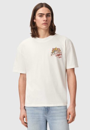 Jeune personne aux cheveux bruns mi-longs portant un t-shirt blanc avec un motif floral coloré et du texte sur le côté supérieur gauche de la poitrine.