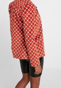 Röd kordjacka med ett vitt diamantmönster, med en avslappnad passform och långa ärmar, kombinerad med svarta vinylshorts.