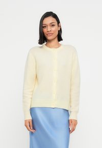 Καρντιγκάν σε ανοιχτό κίτρινο χρώμα, κατασκευασμένο από απαλό ύφασμα, με στρογγυλή λαιμόκοψη, μακριά μανίκια και κούμπωμα με κουμπιά στο μπροστινό μέρος.