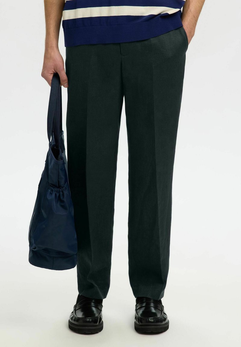 Personne portant un pantalon vert foncé et des mocassins noirs, tenant un sac cabas bleu marine d'une main sur le côté.