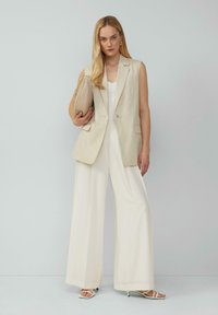 Ärmelloser Blazer aus Leinenmischung in hellbeige über einem weißen Oberteil, kombiniert mit weit geschnittenen cremfarbenen Hosen und weißen Riemchensandalen. Beigefarbene Handtasche inklusive.