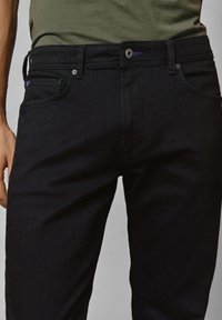 Mies, joka on pukeutunut slim fit -mustiin farkkuihin ja oliivinvihreään paitaan, joissa on näkyvät etutaskut, vyölenkit ja hopeiset napit.