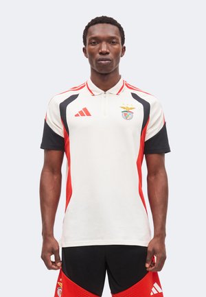 Muž oblečený v bielom, čiernom a červenom polo tričku s logami Benfica a Adidas, v kombinácii s čiernymi a červenými športovými šortkami.