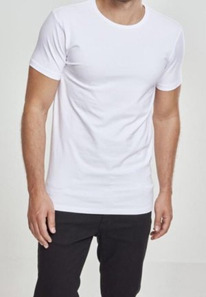 Homme portant un t-shirt blanc uni ajusté à manches courtes et un pantalon noir, debout devant un fond neutre.