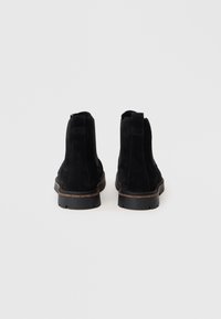 Un paio di stivali neri in suede alla caviglia con suole chunky, mostrati da dietro su uno sfondo bianco semplice.