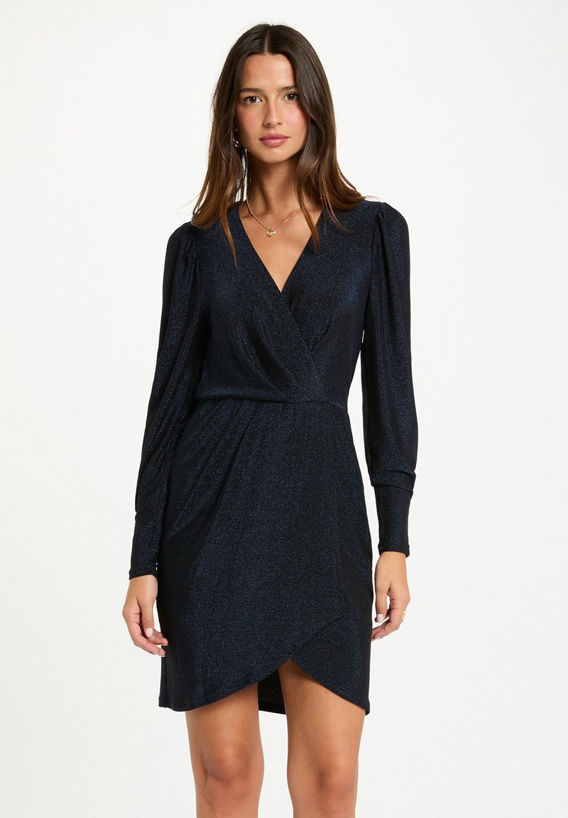 Femme portant une robe noire à manches longues bouffantes et décolleté en V sur fond blanc uni.